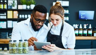 Manager instructs a budtender as they scan a sealed cannabis product label and verify details on a tablet at a dispensary counter, with display cases and a security camera softly blurred in the background.