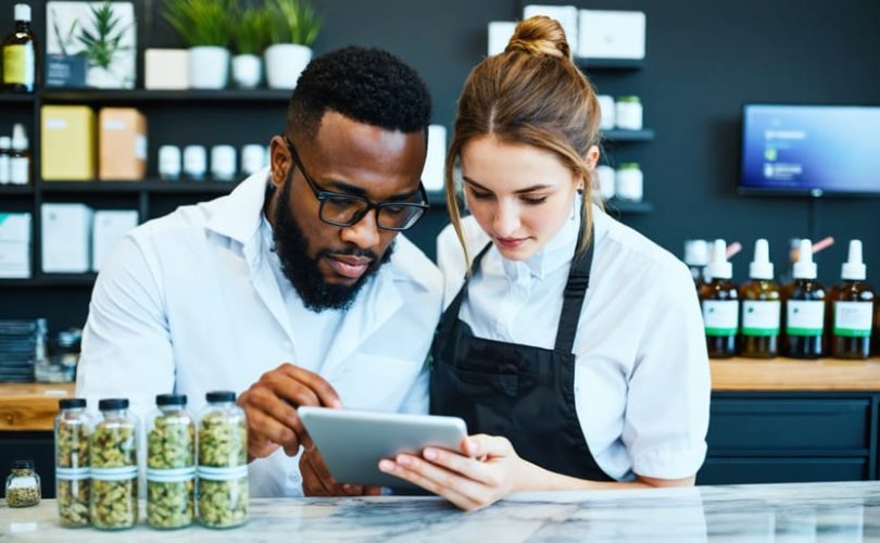 Manager instructs a budtender as they scan a sealed cannabis product label and verify details on a tablet at a dispensary counter, with display cases and a security camera softly blurred in the background.